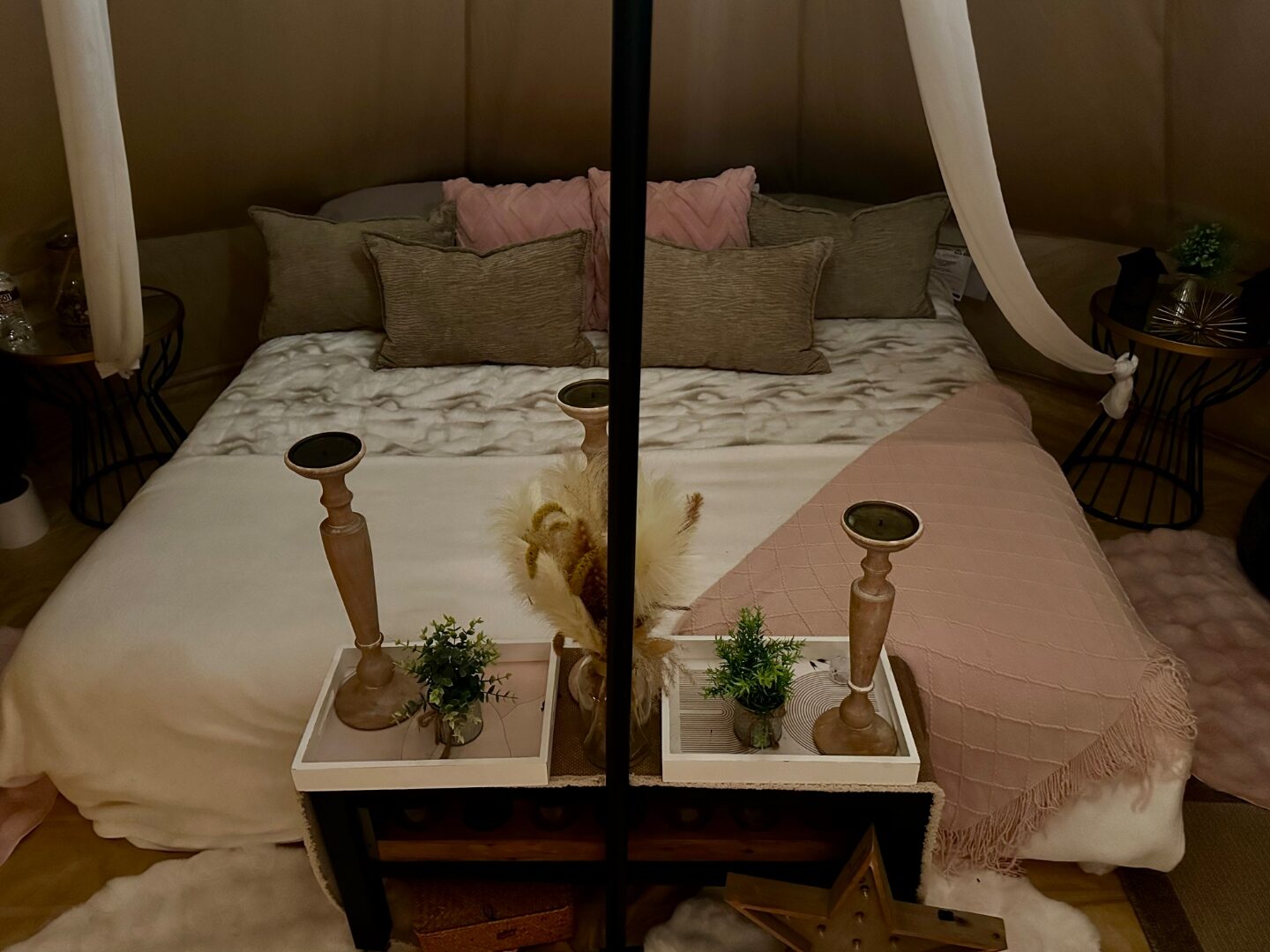 Cama de tienda campana con almohadas rosa y salvia, candelabros de madera y luz suave de linternas.
